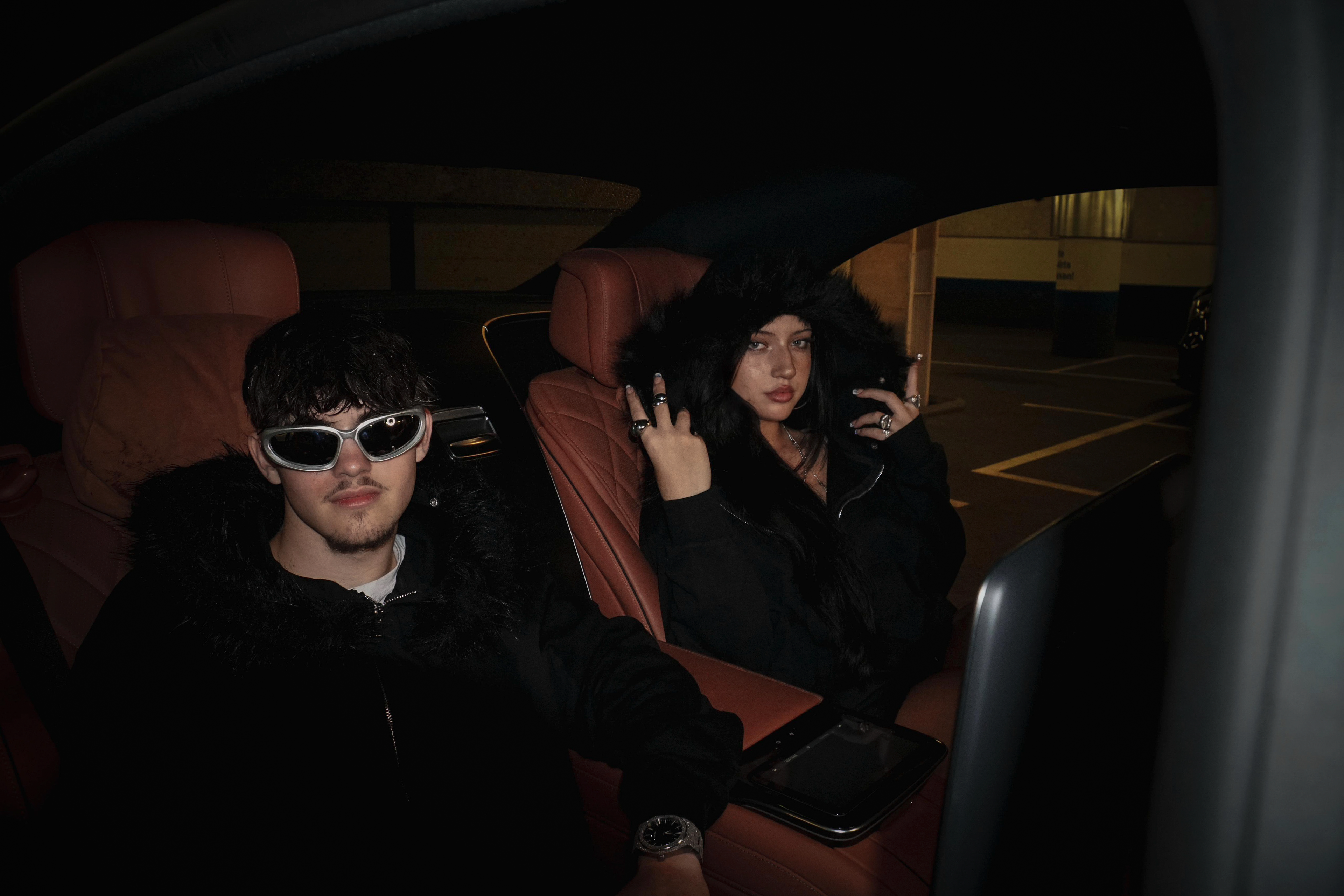 Two people sitting inside a mercedes s class at night, wearing dark clothing and sunglasses.