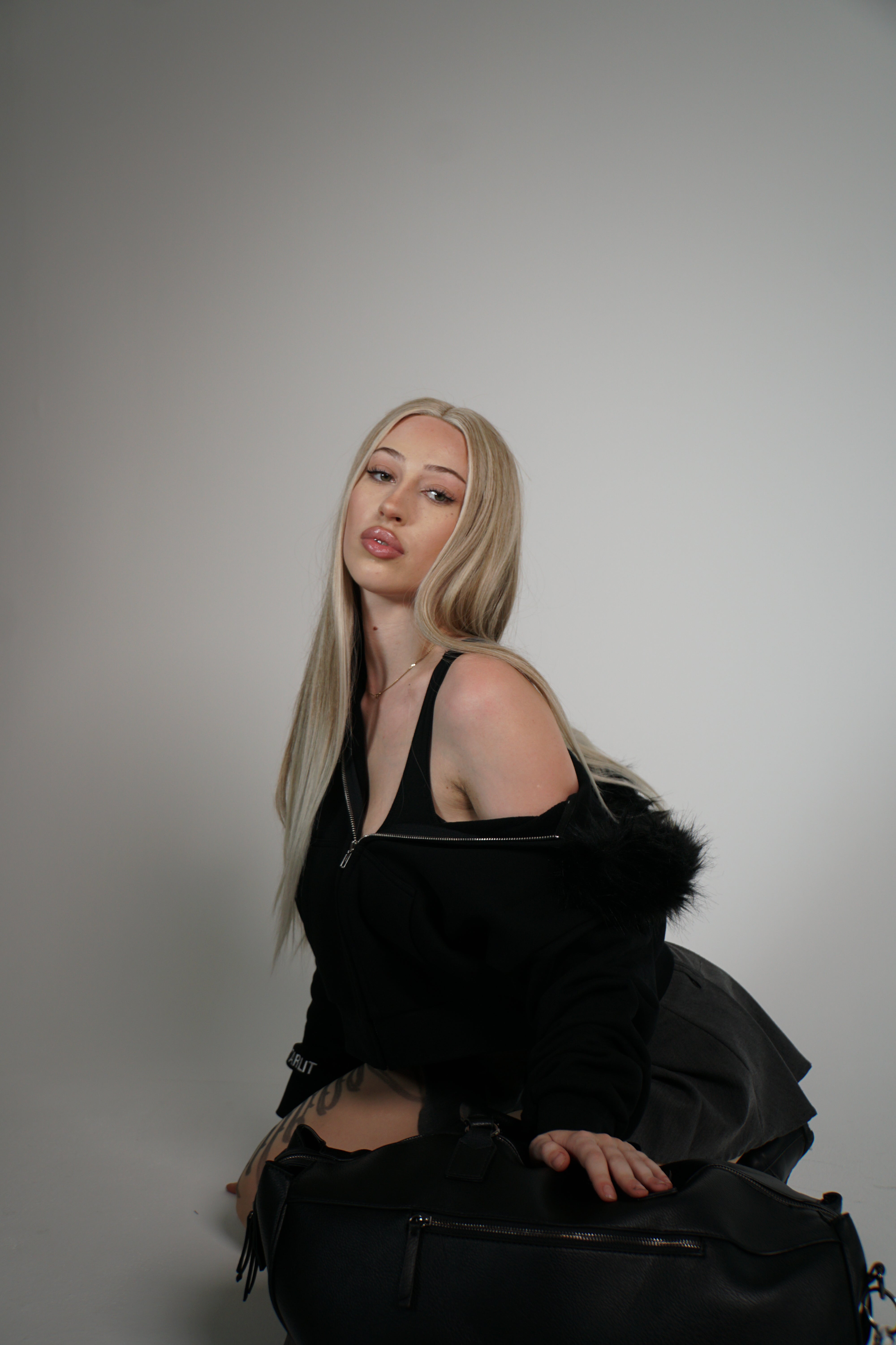 Woman in a black outfit with fur trim sitting on a black bag against a gray background