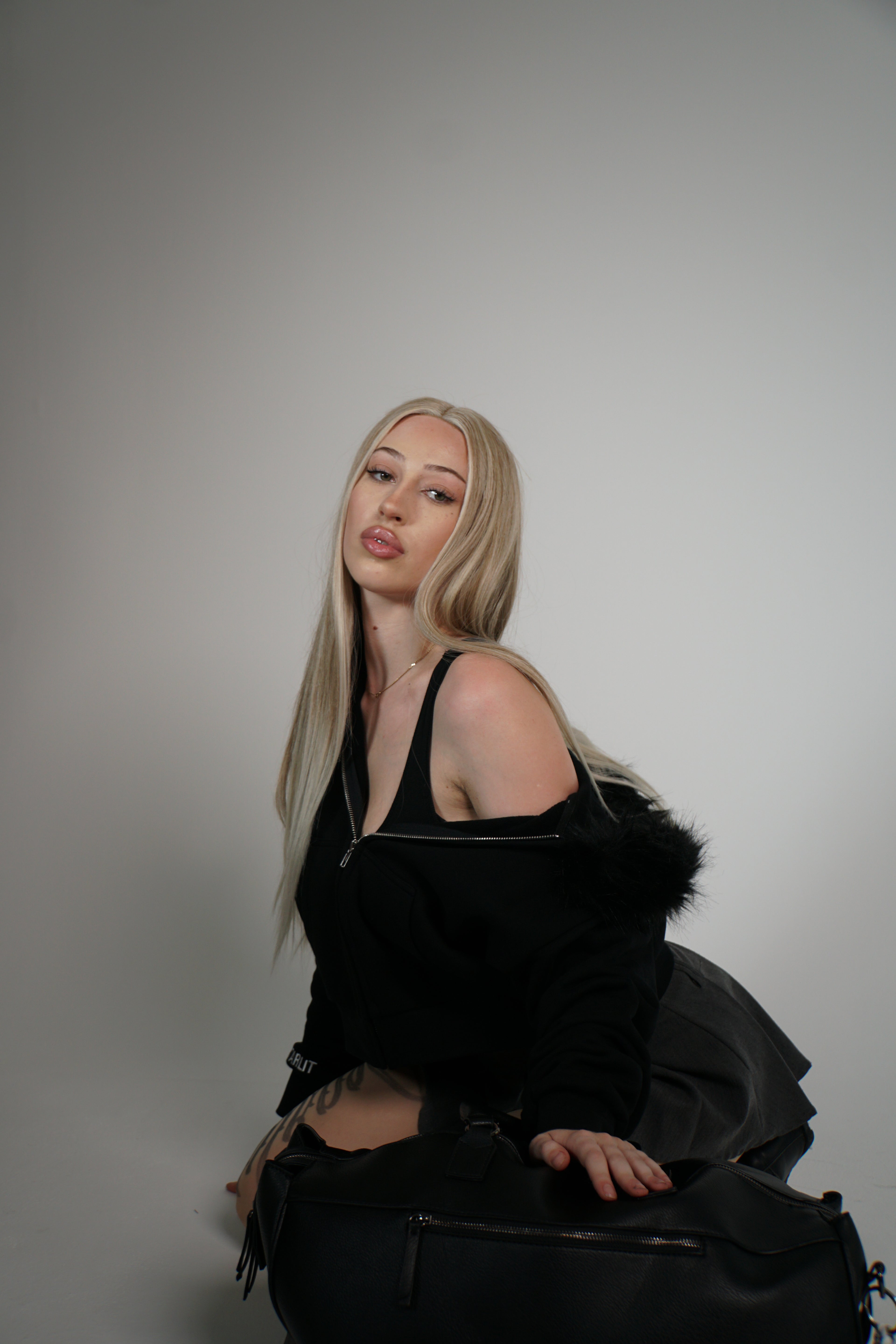 Woman in a black outfit with fur trim sitting on a black bag against a gray background