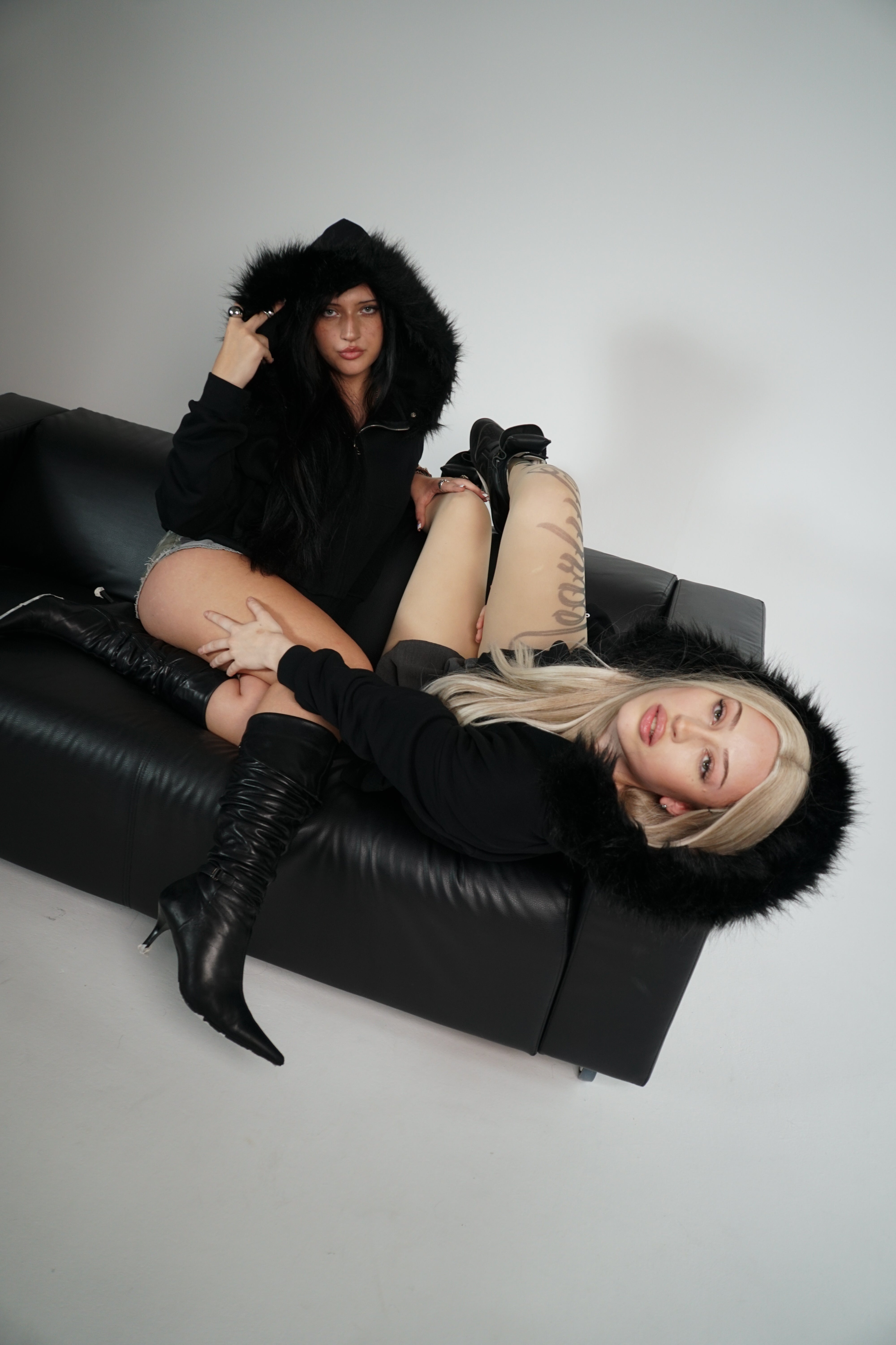Two people wearing black fur-lined hoodies sitting on a black couch against a white background.
