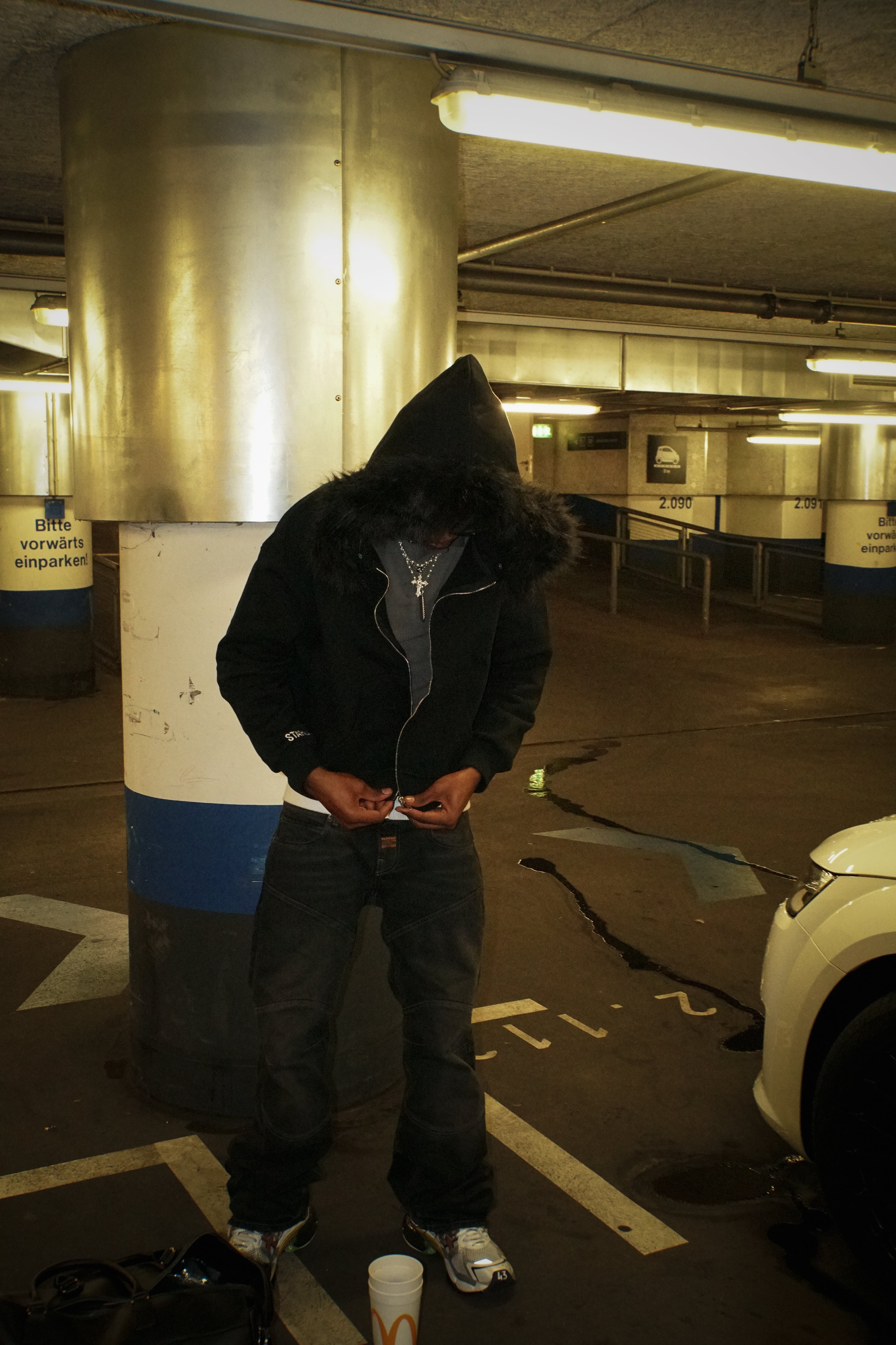 Person in a dark hoodie standing in a parking garage with a cup and phone.