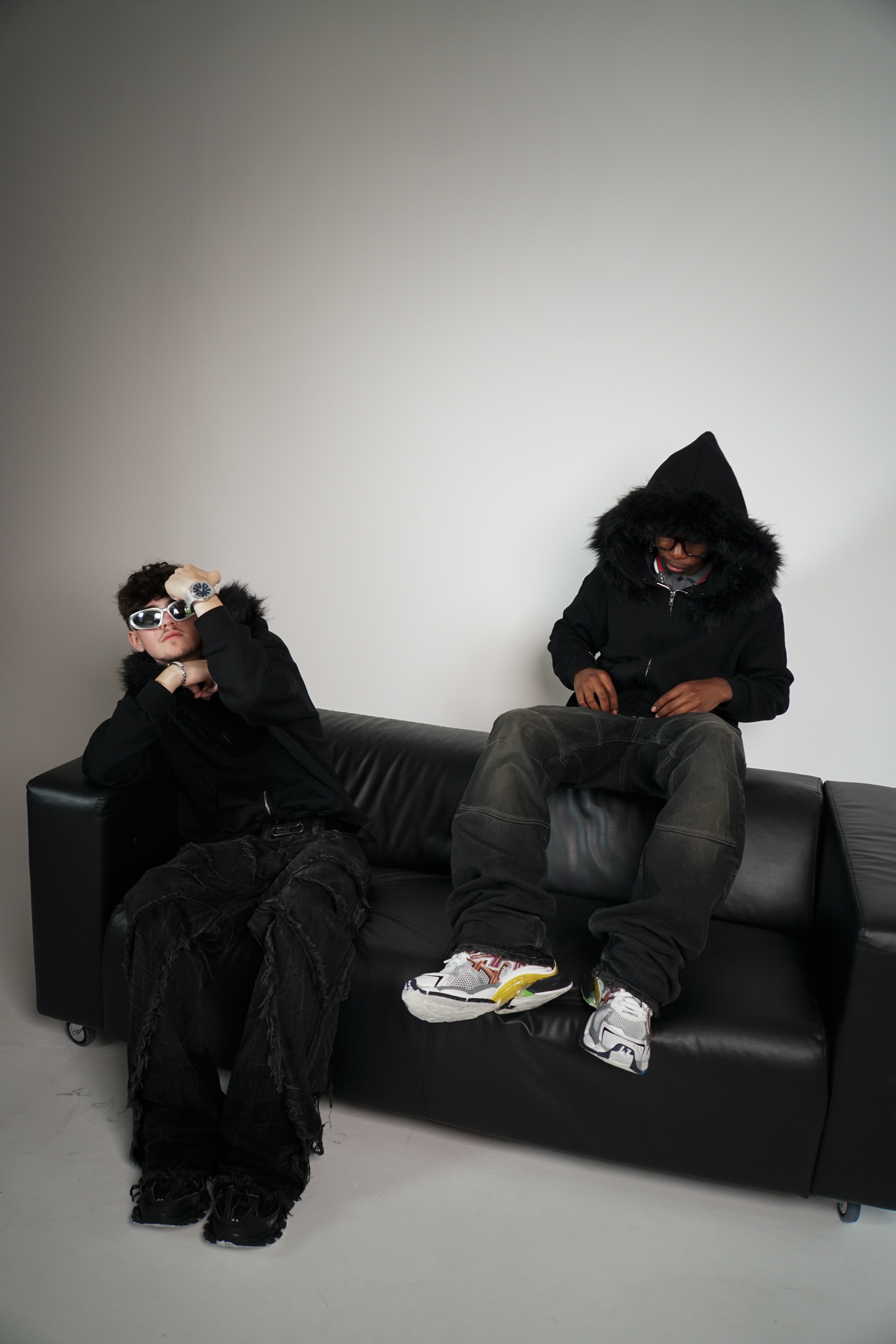 Two people in black hoodies sitting on a black couch against a plain wall.