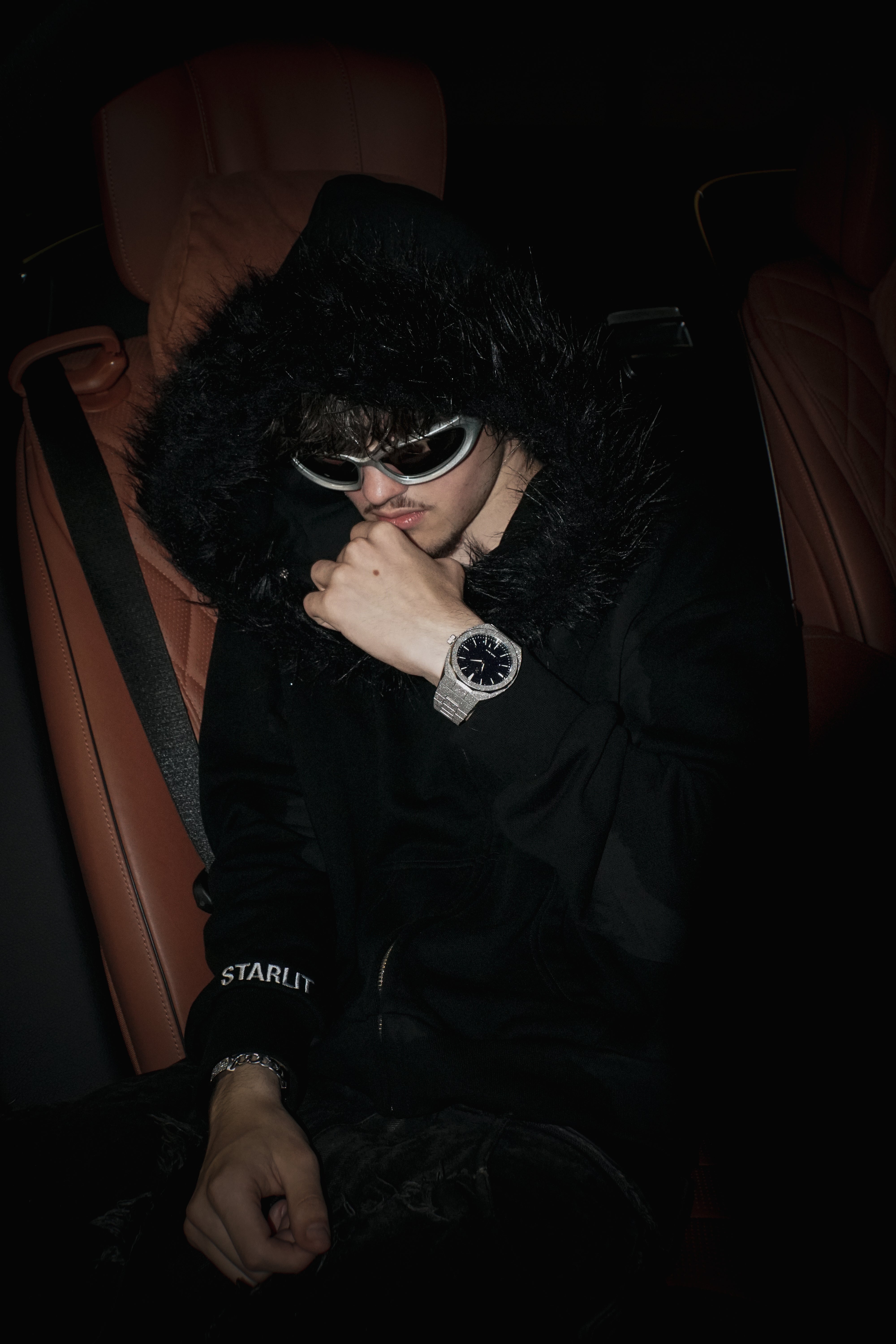 Person wearing a black hoodie with fur lining, sunglasses, and a watch in a dark setting.