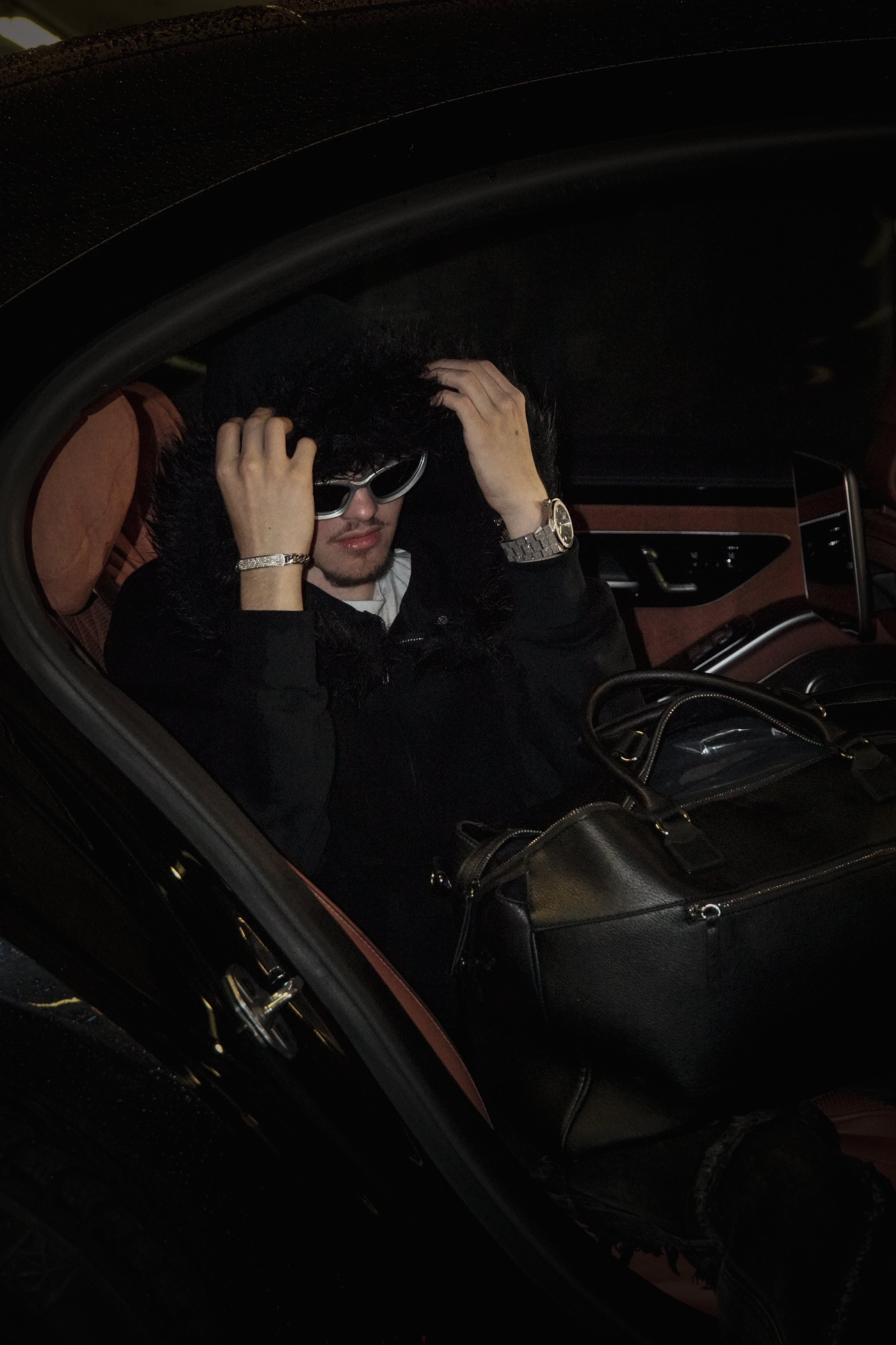 Person sitting inside a car, wearing sunglasses and a dark coat.