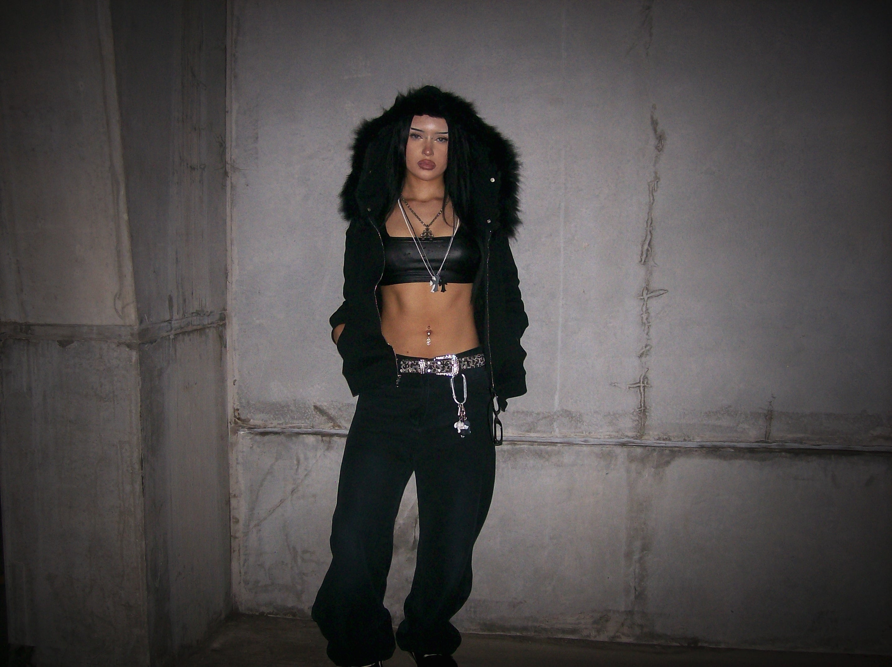 Person wearing a black outfit with a fur-lined hood in a dimly lit room.