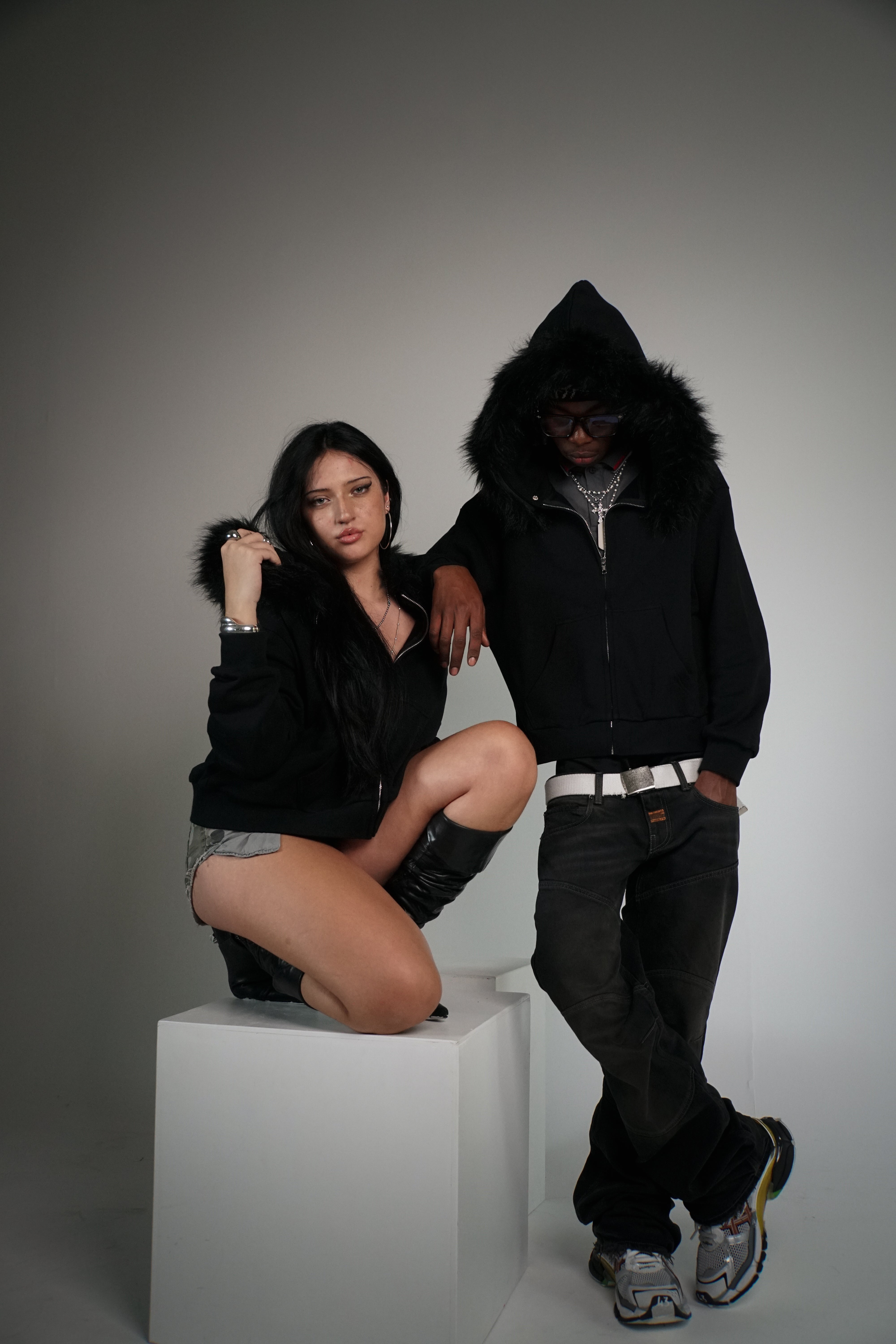 Two people wearing black hoodies with fur-lined hoods on a gray background